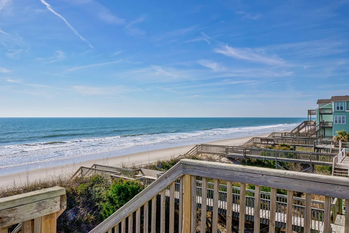 A breathtaking view of the ocean with a sandy beach, wooden walkway, and lush dune vegetation in the foreground. The tranquil setting evokes the peacefulness of coastal living and the allure of direct beach access just steps away from the property.