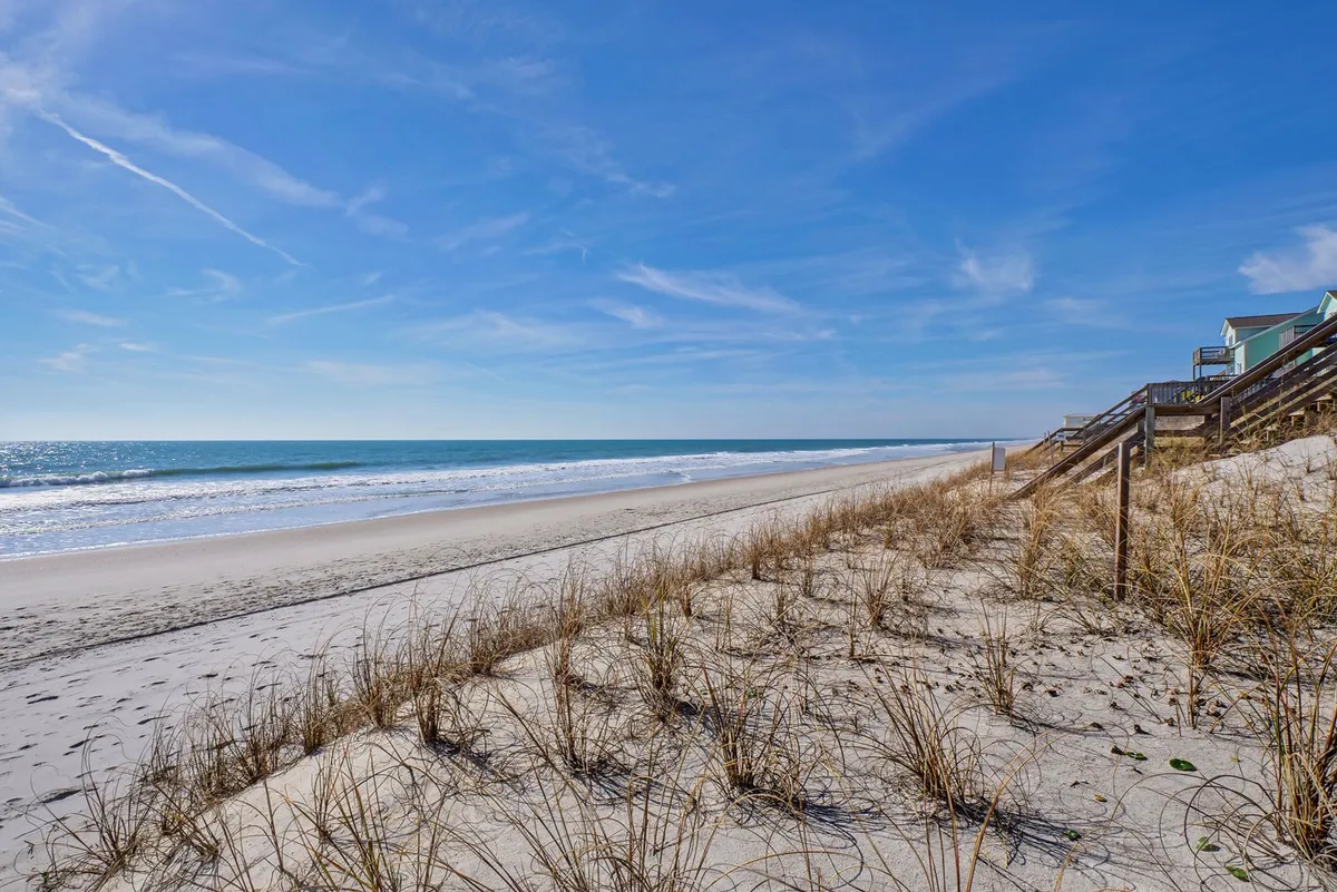 A wide stretch of sandy beach bordered by dune grass, with rolling ocean waves and a tranquil sky overhead. The image evokes a sense of peacefulness and space, ideal for family activities like sandcastle building, strolls, and relaxing by the surf.