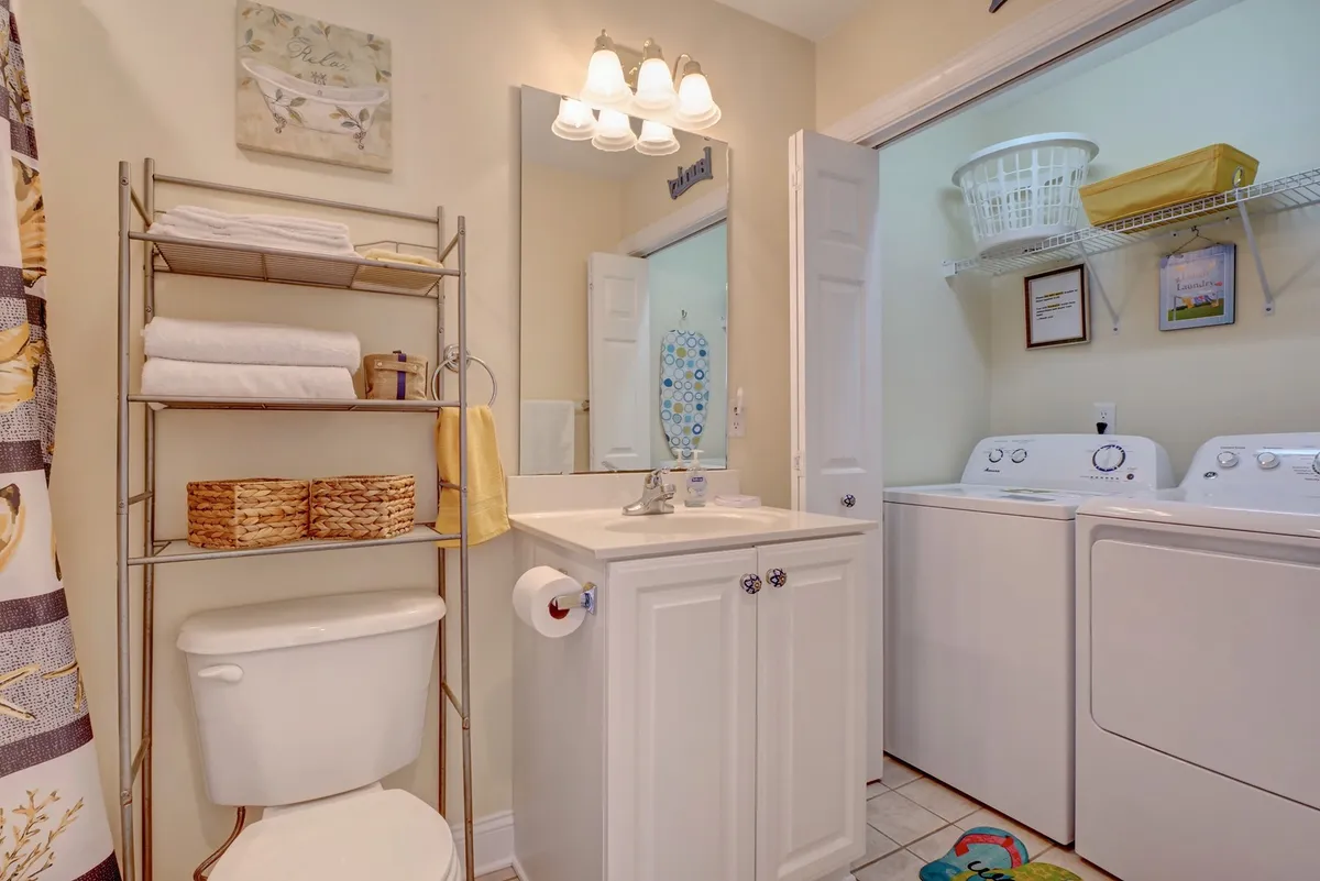 A functional and inviting laundry and bath area featuring a vanity with sink, a stacked washer and dryer, towel rack, toilet, and decorative artwork. The space is practical yet pleasant, streamlining post-beach routines.