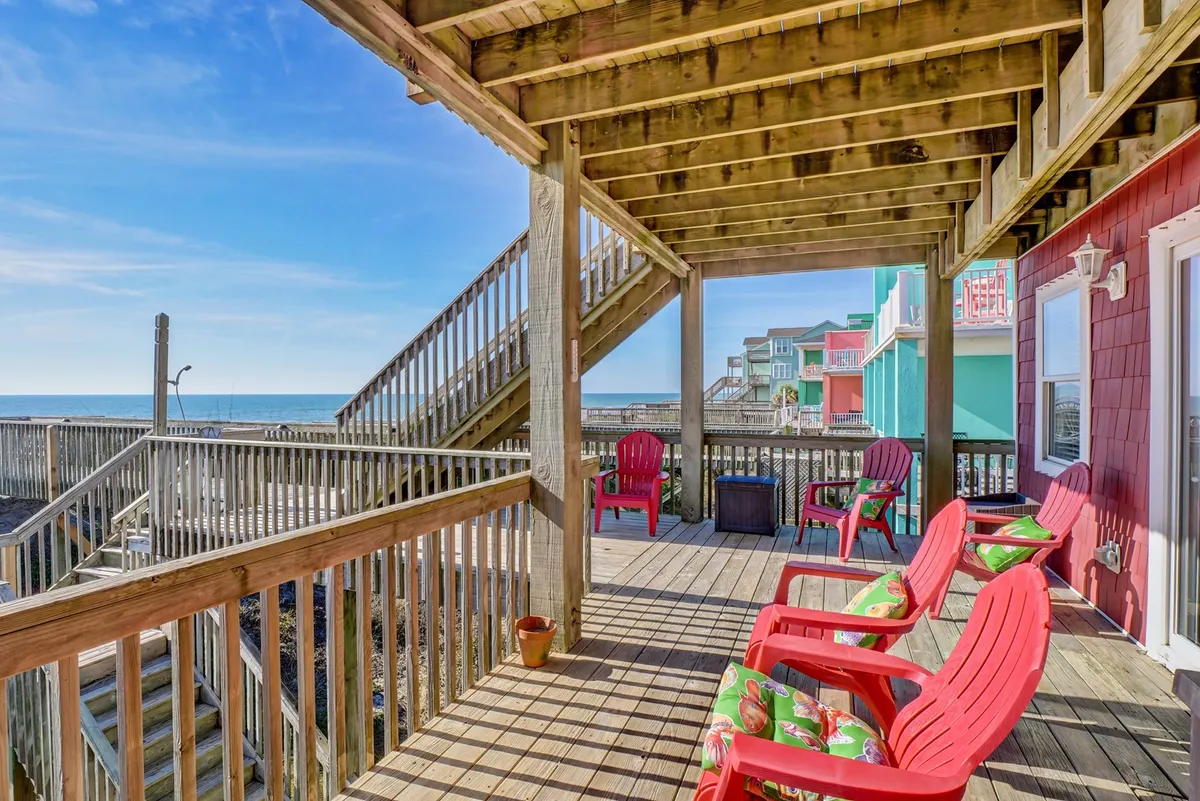 This image features red Adirondack chairs arranged on a wooden deck overlooking the ocean. The staircase leading down from the deck hints at direct beach access, while the expansive horizon suggests both sunrise and sunset views. The deck is furnished and designed for relaxation, socializing, and enjoying the outdoors at any hour.