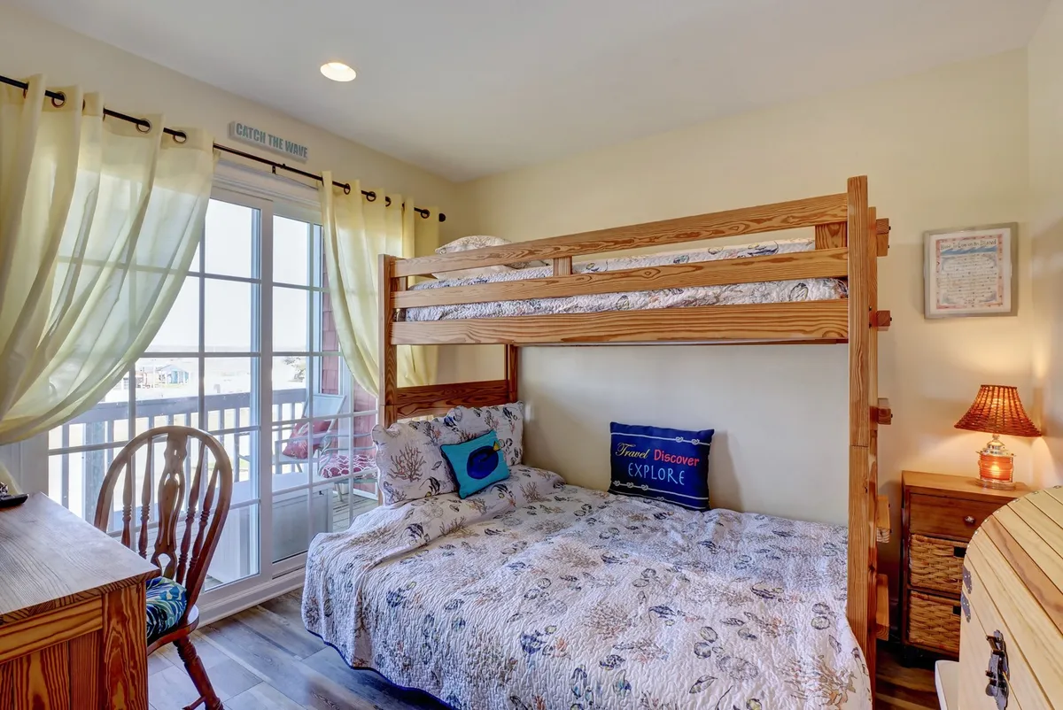 A playful pyramid bunk bedroom with a bunk bed, a blue throw pillow reading 'EXPLORE,' a wooden chair, and a window with curtains letting in natural light. The space feels fun and relaxing—ideal for children or friends to unwind after a day at the beach.