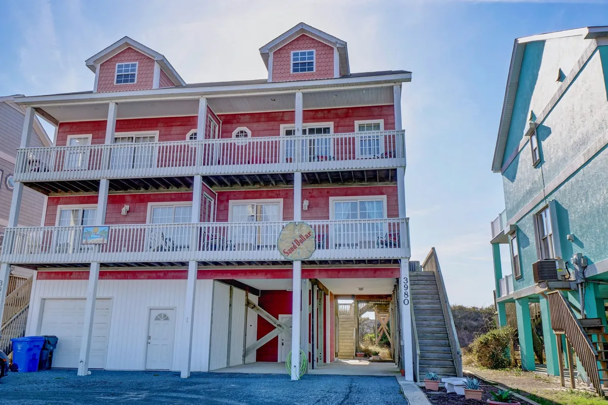 The cheerful red exterior of the house, with multiple balconies and the ‘Sandshack’ sign on display. The covered decks are visible and inviting, suggesting space for family barbecues, games, and ocean viewing—all in a welcoming, vacation-ready setting.