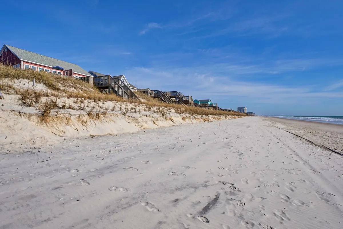 Beachfront homes nestle behind natural sand dunes, with footprints meandering across the soft sand toward the shoreline. The scene is calm and welcoming, suggesting both community and privacy in a picturesque seaside environment.