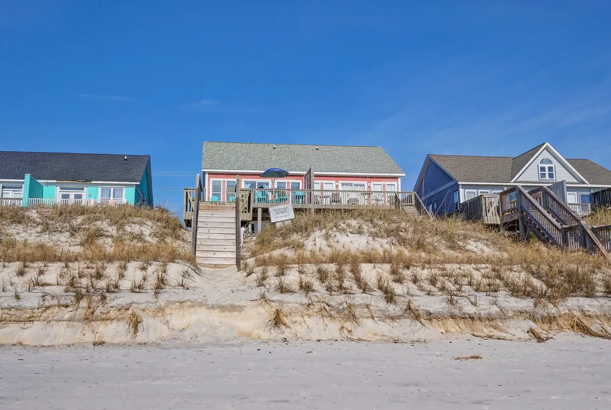 A wide exterior view of the beach house with stairs leading directly down to the sand, surrounded by grass and dunes. The tranquil mood and visible entry emphasize the home’s direct connection to the shoreline.
