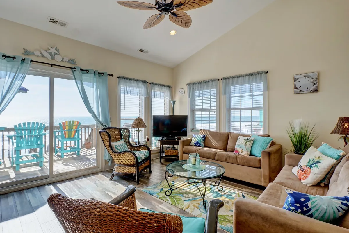 This image features a spacious, open living room with comfortable sofas, colorful cushions, a decorative rug, and large windows that frame an inviting balcony view of the ocean. Natural light floods the space, making it feel airy and bright, while the arrangement of furniture encourages socializing, relaxing, and enjoying the views.