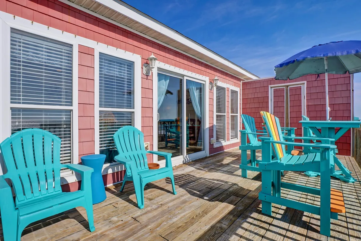 A vibrant outdoor deck featuring colorful Adirondack chairs, bright patio furniture, and a large umbrella, all set next to sliding glass doors that hint at the connection between inside and out. The scene is inviting and lively, perfect for casual relaxation or gathering with a loved one to enjoy the fresh coastal air.