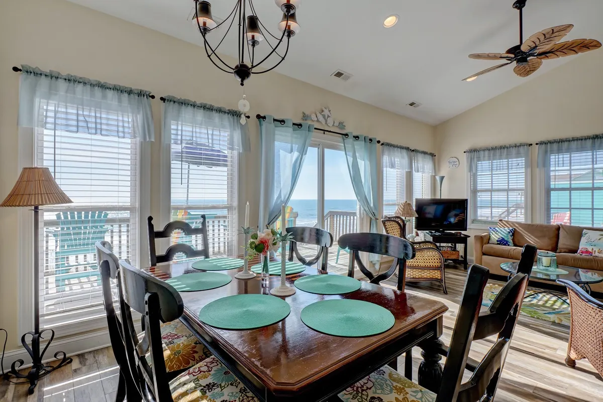 A cheerful dining area with a table set with turquoise placemats, positioned beside expansive windows that showcase a stunning ocean view. The adjacent living space features a comfortable couch, and a decorative chandelier hangs overhead, creating a bright, inviting atmosphere for family meals or casual gatherings.