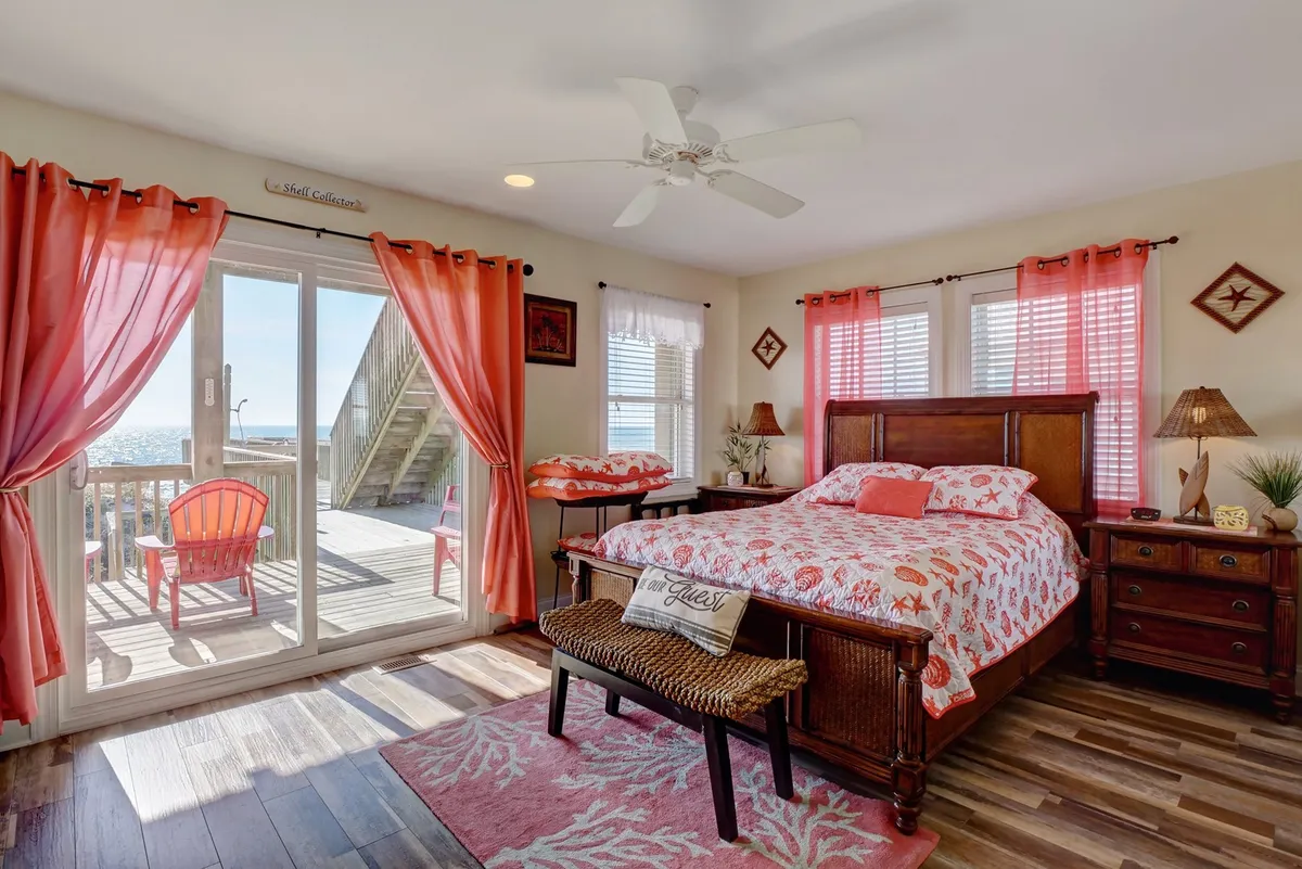 A cheerful master suite with coral-patterned bedding, a sliding glass door that opens onto a deck with red Adirondack chairs, and colorful curtains. Sunlight fills the room, creating a bright and welcoming atmosphere perfect for relaxing or enjoying coastal views from bed.