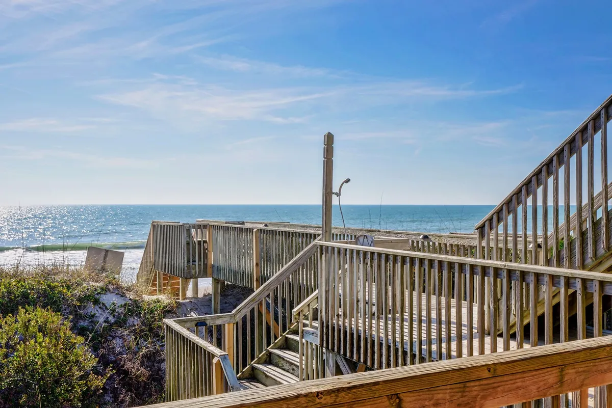 A wooden boardwalk leads directly from the house over dune vegetation to a stunning, open ocean view. The scene captures the anticipation of stepping out from your accommodation and immediately connecting with the beach environment, with soft sand and calm sea waves visible ahead under a relaxed sky.