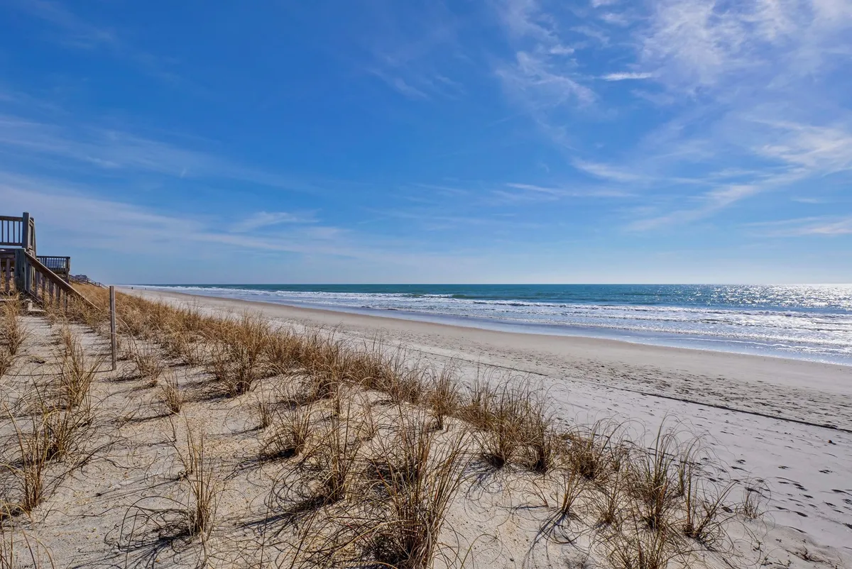 A sweeping view of the beach with golden sand, rolling dune grass, a classic boardwalk, and the glistening ocean stretching into the horizon. There's a gentle, relaxing vibe, with traces of footprints in the sand and the early morning sun illuminating the scene.
