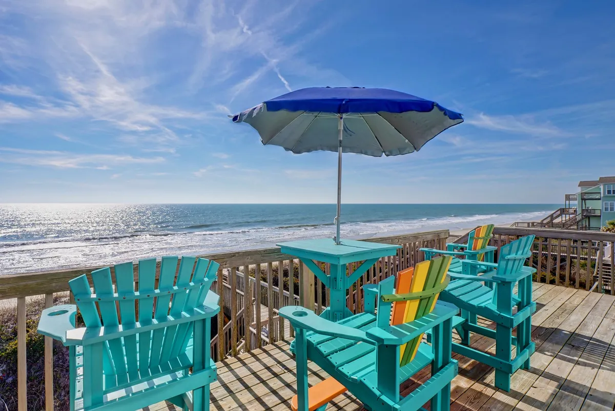 The image presents the exterior of an oceanfront duplex at North Topsail Beach, showcasing a row of vibrant Adirondack chairs under a shady umbrella, all facing a sweeping, unobstructed view of the Atlantic. The setup invites relaxation, conversation, and sunset watching, with the tranquil ocean stretching beyond.