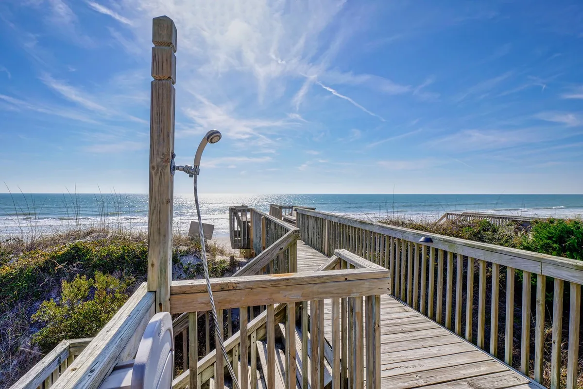 A serene exterior view featuring a wooden walkway leading directly to the beach, with an enclosed outdoor shower visible to the side, set against dunes and the ocean beyond. The pathway has benches for seating, making it a welcoming space for pre- and post-beach moments.