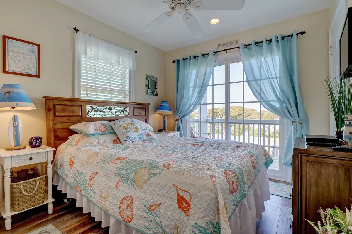A cozy bedroom featuring a bed with a colorful quilt, large window with sheer curtains allowing in natural light, a nightstand with a lamp, and decorative wall art. The room feels tranquil and inviting, ideal for relaxation and peaceful mornings.